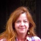 Eine Frau in einem karierten Hemd lächelt am Eingang eines Hauses mit einer Willkommensnachricht und einem lustigen Spruch.