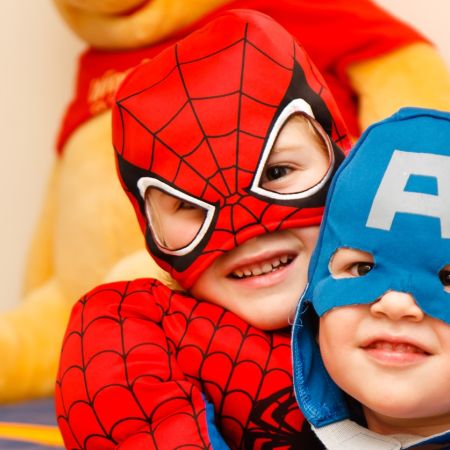 Zwei kleine Kinder in Superheldenkostümen, Spiderman und Captain America, lächeln fröhlich zusammen im Zimmer.