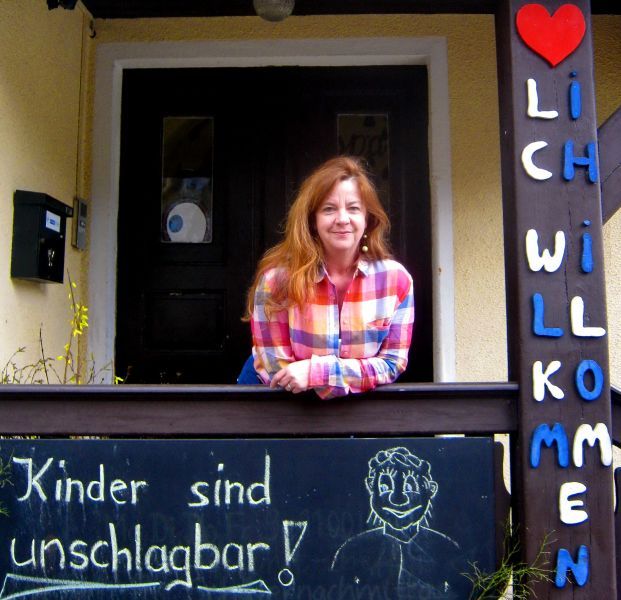 Eine Frau in einem karierten Hemd lächelt am Eingang eines Hauses mit einer Willkommensnachricht und einem lustigen Spruch.